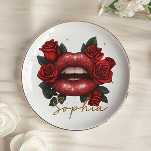Könnte beinhalten: Weiße Keramikplatte mit goldenem Rand, verziert mit roten Lippen, umgeben von roten Rosen und grünen Blättern. Der Name "Sophia" ist in goldener Schrift darunter geschrieben.