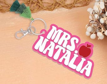 May include: A pink keychain with the text "MRS NATALIA" in white, with a red apple graphic. The keychain has a silver clasp and ring, and a green tassel. The background is a light peach color.