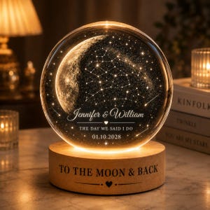 May include: A spherical glass night light featuring an etched moon and constellation design. The glass sphere sits on a cylindrical wooden base engraved with the text TO THE MOON & BACK. The sphere displays Jennifer & William, THE DAY WE SAID I DO, and 01.10.2028.