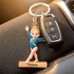 May include: A keychain featuring a gymnast figurine in a blue leotard with the text "2025" on a green sash. The figurine is standing on a wooden base with the name "Victoria" printed on it. A car key fob is next to the keychain.