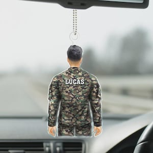 May include: A car hanging ornament featuring a soldier in a camouflage uniform. The ornament is a cutout of a person from the back, with the name "LUCAS" printed on the back of the uniform. The ornament hangs from a silver chain.