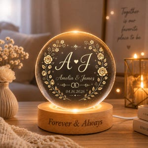 May include: A circular glass plaque illuminated by a wooden base engraved with the text Forever & Always. The glass features an etched floral wreath surrounding the initials A & J, the names Amelia & James, two rings, and the date 04.16.2026.