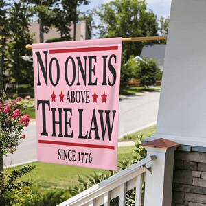 May include: A pink house flag with the words "NO ONE IS ABOVE THE LAW" in black, with red stars and a red banner that reads "SINCE 1776". The flag is hanging on a wooden pole on a porch.