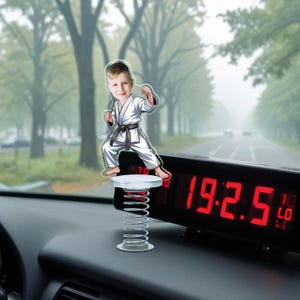 May include: A cartoon figure of a young boy in a karate uniform, with a black belt, is mounted on a spring. The figure is positioned in front of a digital clock displaying red numbers. The background shows a road lined with trees.