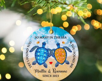 May include: Round ceramic ornament featuring two sea turtles, one blue and one orange, with a heart between them. Text includes "SO MANY IN THE SEA", "YET I FOUND YOU", "& YOU FOUND ME", "Mattie & Roxanne SINCE 2023". The ornament hangs from a gold string.