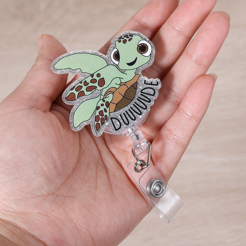 May include: A badge reel featuring a cartoon sea turtle with the word "DUUUUDE". The turtle has a green body, brown shell, and brown spots on its flippers. The reel has a clear plastic strap and a metal clip.