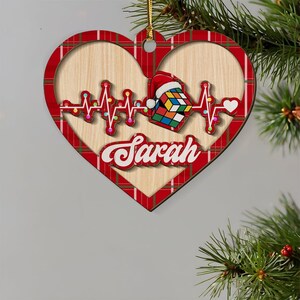 May include: Heart-shaped wooden Christmas ornament with a red plaid border. The ornament features a Rubik's Cube wearing a Santa hat, a heartbeat design, and the name "Sarah". The ornament is hanging from a gold string.