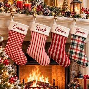 Puede incluir: Cuatro calcetines de Navidad colgados de una repisa, cada uno con un puño de felpa blanca bordado con los nombres Emma, Noah, Olivia y James. Los calcetines presentan patrones de cuadros vichy, rayas verticales rojas y blancas, y cuadros escoceses.
