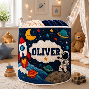 May include: A cylindrical fabric storage basket with a quilted dark blue exterior featuring a space-themed design. The basket displays a rocket, planets, a moon, and an astronaut on a moon surface, with the name OLIVER centered in a white cloud.