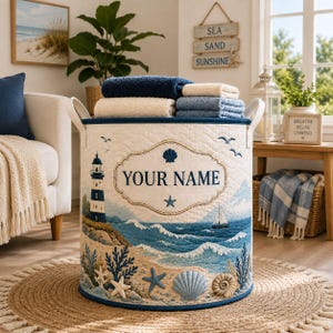 May include: A cylindrical fabric storage basket with quilted white material featuring a coastal scene with a lighthouse, ocean waves, seashells, and the text YOUR NAME. The basket has two side handles and contains folded blue and white towels.