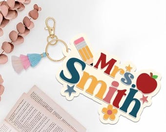 May include: A colorful keychain with the name "Mrs. Smith" in a playful font, featuring a pencil, apple, and flower. The keychain has a gold clasp and a tassel with pink, blue, and white accents. A book is in the background.