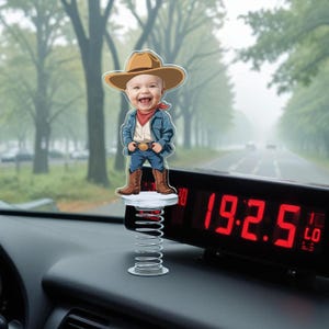 May include: A novelty dashboard bobblehead featuring a smiling baby dressed as a cowboy. The figure wears a brown cowboy hat, blue jacket, jeans, and boots. The bobblehead is mounted on a silver spring.