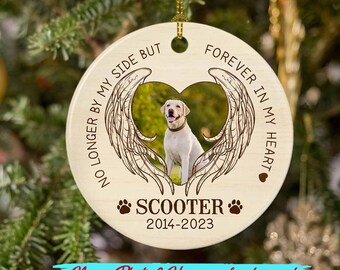 Puede incluir: Un adorno navideño redondo de color crema con una foto de un perro dentro de un marco en forma de corazón con alas de ángel. El adorno tiene el texto "NO LONGER BY MY SIDE BUT FOREVER IN MY HEART" y el nombre "SCOOTER 2014-2023". También está presente el texto "Name Photo & Year can be changed".