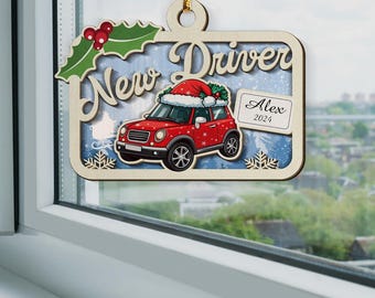 Pode incluir: Enfeite de Natal de madeira com um carro vermelho com um chapéu de Papai Noel, as palavras "New Driver" e "Alex 2024". O enfeite tem um design de azevinho e bagas e detalhes de flocos de neve, pendurado por um fio dourado.