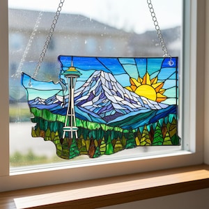May include: Stained glass art piece in the shape of Washington state, featuring the Space Needle, a mountain range, and a yellow sun. The artwork is framed with a silver chain for hanging. The colors include blue, green, yellow, and white.