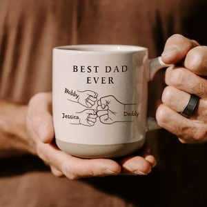 May include: A cream-coloured ceramic mug with a brown base and a brown printed design. The text reads BEST DAD EVER above a graphic of three fists bumping together. The names Bobby and Jessica are printed near the smaller fists, and Daddy is printed near the larger fist.