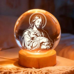 Puede incluir: Una esfera de vidrio transparente con una imagen grabada de Jesús sosteniendo un cordero, colocada sobre una base de madera. La imagen está iluminada desde adentro, creando un brillo cálido. La esfera tiene un halo alrededor de la cabeza de Jesús.