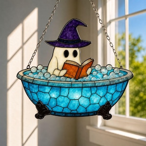 Op de afbeelding: Een glas-in-lood suncatcher met een witte geest die een paarse heksenhoed draagt, zittend in een blauw bad op pootjes gevuld met heldere glazen bubbels terwijl hij een open oranje boek leest. Het stuk hangt aan een zilveren metalen ketting.