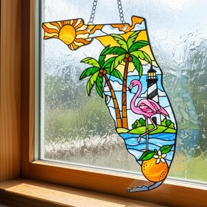 May include: A stained glass window hanging in the shape of Florida, featuring a sunny sky, palm trees, a pink flamingo, and a lighthouse. An orange with a flower is at the bottom. The window is suspended by a silver chain.