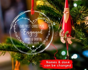 May include: Clear glass Christmas ornament with the words "OUR FIRST CHRISTMAS ENGAGED" and the names "AMIRI & GRIFFIN" along with the date "05.12.2024" in white script. The ornament is heart-shaped and hangs from a red ribbon. A red Santa Claus ornament is also visible.