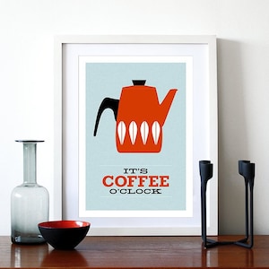 May include: A framed print with a retro coffee pot design in orange, black, and white on a light blue background. The text "IT'S COFFEE O'CLOCK" is below the image. A glass vase, a black and red bowl, and black candle holders are on a wooden surface.