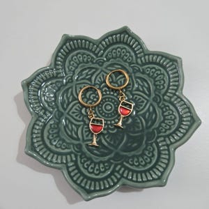 May include: A pair of gold hoop earrings with a red wine glass charm hanging from each hoop. The earrings are on a green mandala-patterned plate.