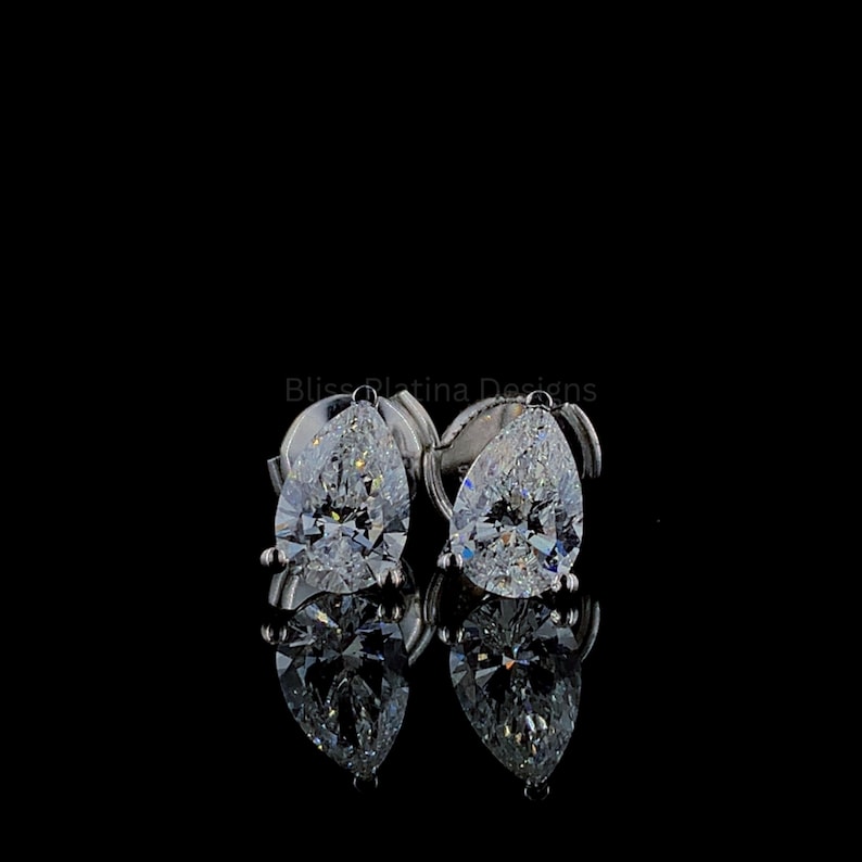 Pu&ograve; includere: Un paio di orecchini a lobo con taglio a goccia caratterizzati da pietre trasparenti sfaccettate incastonate in metallo color argento lucido. Gli orecchini sono posizionati su uno sfondo nero con il testo Bliss Platina Designs visibile sopra i gioielli.