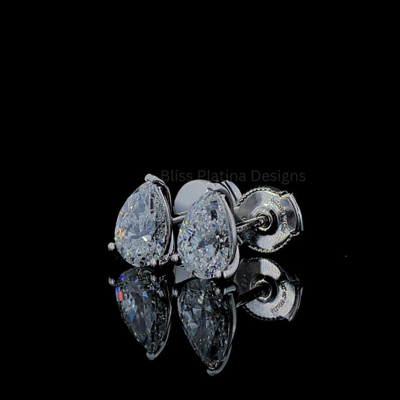 Pu&ograve; includere: Un paio di orecchini a lobo a forma di pera con pietre sfaccettate trasparenti incastonate in metallo color argento lucido. Gli orecchini includono chiusure a vite e sono mostrati su uno sfondo nero con il testo Bliss Platina Designs.