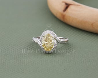 May include: A silver-toned ring featuring a central oval-cut yellow gemstone surrounded by a halo of small white stones. The band has a bypass design with pavé-set white stones along the shoulders. The text Bliss Plated Designs is visible.