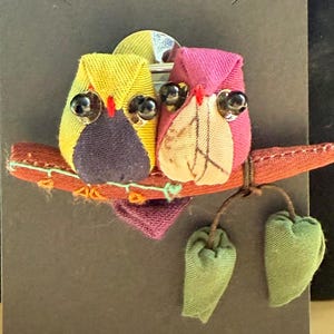 May include: A fabric brooch featuring two stylised owls perched on a branch. The left owl is yellow and purple, while the right owl is pink and beige. Both have black bead eyes and red stitched beaks. Two green fabric leaves hang from the branch.