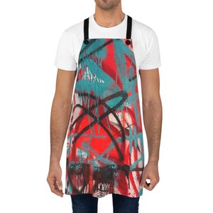May include: An apron featuring a vibrant graffiti-style print with overlapping layers of bright red, teal, and black spray paint patterns. The design includes drips, splatters, and bold black lines, finished with solid black neck and waist straps.