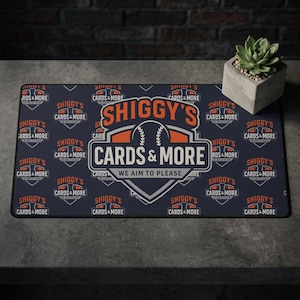 以下が含まれることがあります： 白とオレンジ色の「SHIGGY'S CARDS & MORE」の文字が入った長方形の濃い青色のマット。マットには野球のグラフィックと「WE AIM TO PLEASE」というフレーズが描かれています。コンクリートの鉢に入った小さな多肉植物が右側にあります。