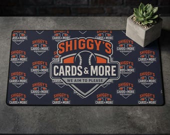 Peut inclure: Un tapis rectangulaire bleu marine avec le texte "SHIGGY'S CARDS & MORE" en blanc et orange. Le tapis présente un graphisme de baseball et la phrase "WE AIM TO PLEASE". Une petite plante succulente dans un pot en béton est à droite.