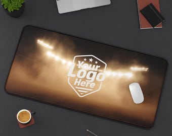 Peut inclure: Tapis de souris rectangulaire avec un motif de projecteurs de stade et le texte "Your Logo Here" dans un bouclier blanc. Une souris blanche, un ordinateur portable et d'autres fournitures de bureau sont sur une surface gris foncé.