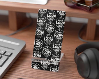 Peut inclure: Un support de téléphone noir avec un motif de logo blanc répété qui dit "Your Logo Here". Le support est en métal et tient un téléphone. L'arrière-plan comprend un clavier, un casque et un écran d'ordinateur.