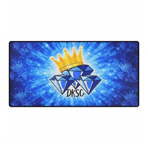 May include: A rectangular desk mat featuring a blue background with a diamond and crown design. The design includes three blue diamonds, a gold crown, and the text "DKSC". The mat has a blue and white starburst pattern.