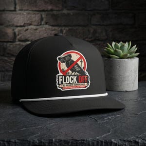 May include: A black baseball cap with a white cord across the brim. The front features a graphic of a bird perched on a surveillance camera inside a red prohibition circle. Text reads: FLOCK OFF, NOT YOUR DATA, NO PRIVATE SURVEILLANCE.
