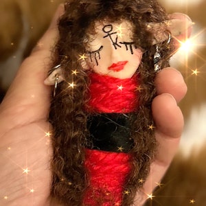 Puede incluir: Una muñeca pequeña con cabello largo y castaño, ropa roja y negra, y una cara pintada con los ojos cerrados y una boca roja. La muñeca tiene un símbolo dibujado en la frente.