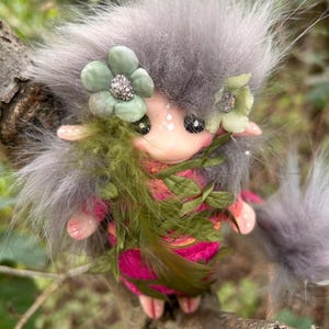 May include: A handmade fairy doll with fluffy grey hair, decorated with green flower accents and a pink and green dress. The doll has a delicate face with black eyes and is embellished with green feathers.