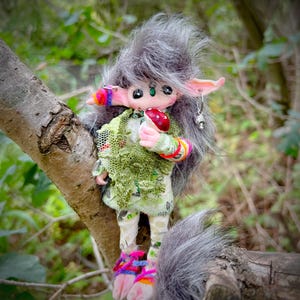 May include: A handmade doll with grey fur, pink ears, and a green outfit. The doll is holding a red mushroom and is adorned with colourful bracelets and anklets. It is perched on a tree branch, with a blurred forest background.
