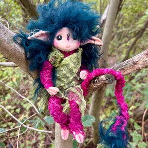 May include: A fantasy doll with a blue, fluffy hairstyle and pointed ears, wearing a magenta outfit with a green overlay. The doll is perched on a tree branch, with a matching magenta and blue tail.