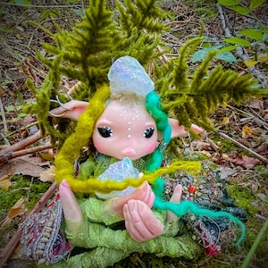 May include: A fairy doll with a pale complexion, large eyes, and pink lips. The doll has green and yellow yarn hair, wears a green dress, and holds a crystal. The background features moss and foliage, creating a woodland theme.