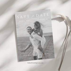 Puede incluir: Tarjeta de guardar la fecha en blanco y negro con una pareja en la playa. El texto dice "Save the Date" con la fecha "July 15, 2023" y la ubicaci&oacute;n "Newport, RI". Los nombres de la pareja est&aacute;n escritos en cursiva debajo de la foto: "amber & brian".