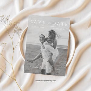 Puede incluir: Una tarjeta de guardar la fecha en blanco y negro con una pareja en la playa. El texto dice "Save the Date" con la fecha "July 15, 2023" y la ubicaci&oacute;n "Newport, RI". Los nombres de la pareja son "Amber & Brian".
