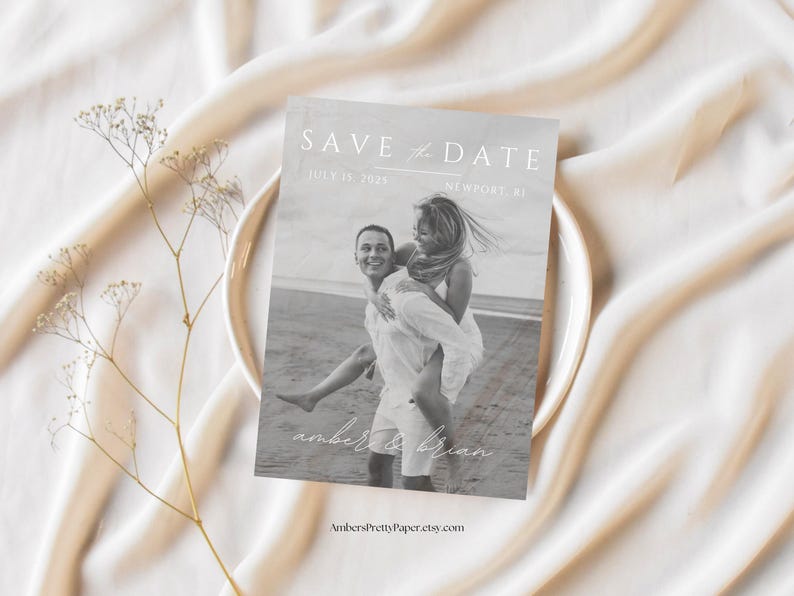 Puede incluir: Una tarjeta de guardar la fecha en blanco y negro con una pareja en la playa. El texto dice "Save the Date" con la fecha "July 15, 2023" y la ubicaci&oacute;n "Newport, RI". Los nombres de la pareja son "Amber & Brian".