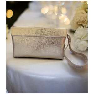 May include: A metallic gold clutch with a textured surface and a wrist strap. The word "ALULA" is subtly embossed on the flap. The clutch is rectangular and appears to be made of leather or a leather-like material.