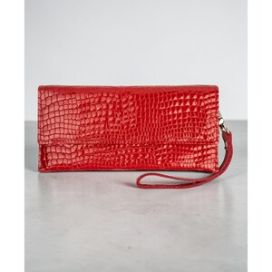 May include: A red, textured clutch with a wrist strap. The clutch has a glossy finish and a rectangular shape with a fold-over flap. The material appears to mimic the look of crocodile skin. The wrist strap is attached to the side with a silver-colored metal clasp.