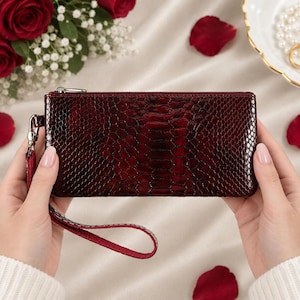 May include: A rectangular wristlet clutch in a deep burgundy color with a glossy, textured snakeskin-like pattern. The accessory features a silver-toned metal zipper closure along the top edge and a matching burgundy wrist strap attached to the side.
