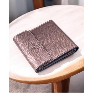 May include: A brown leather wallet with a textured finish. The rectangular wallet has a flap closure and a darker interior. It rests on a light-coloured wooden table.