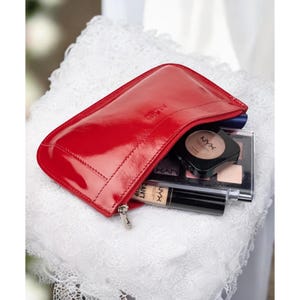 May include: A red leather cosmetic bag with a silver zipper, partially open to reveal makeup products. The bag is on a white lace surface. Makeup products include a compact and a tube of concealer.